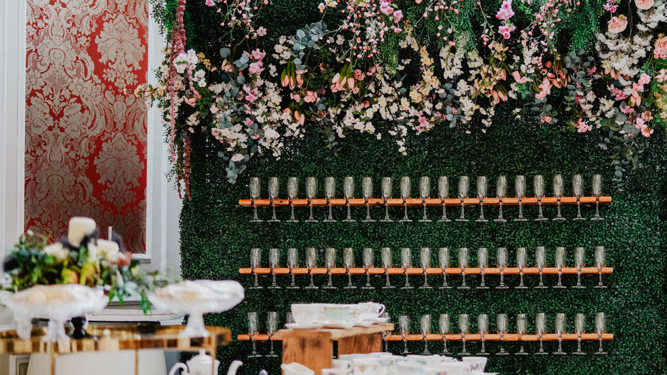 Elegant floral wall with hanging glasses, opulent red damask, and luxurious party setting ready for guests.