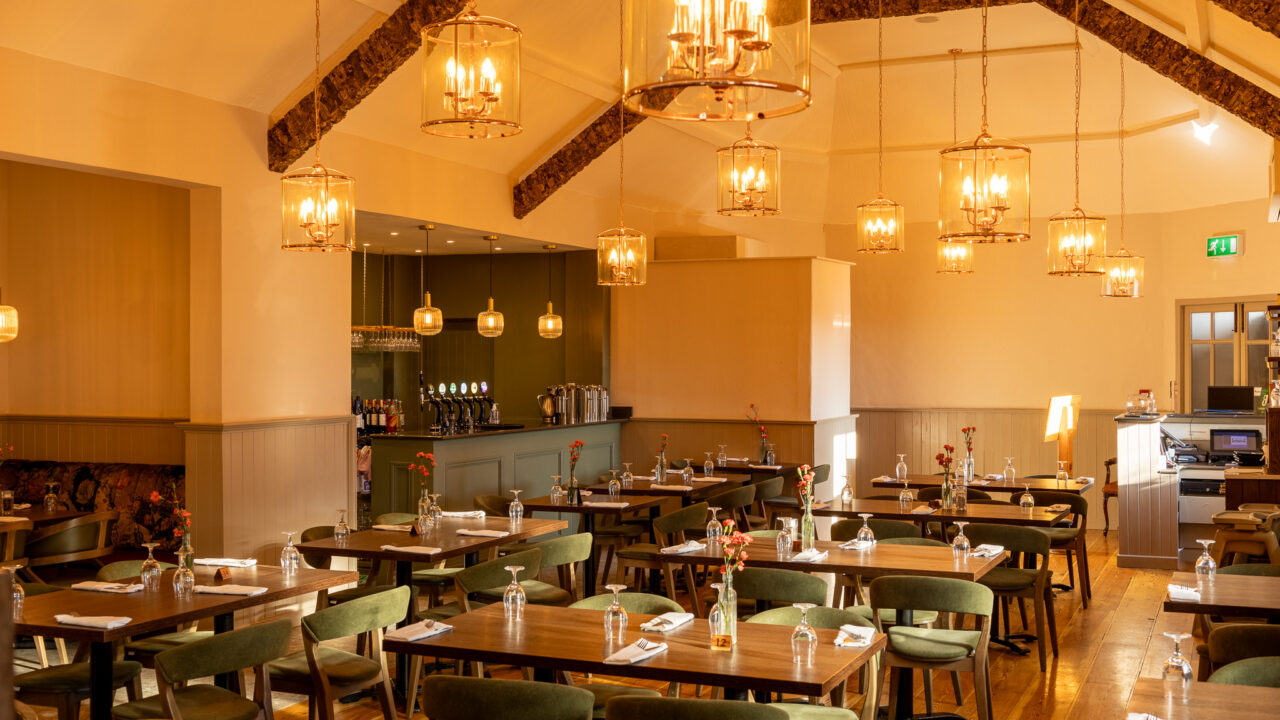 Cosy hotel dining area with rustic chandeliers, wooden tables, elegant green chairs, and a charming bar, perfect for relaxation.