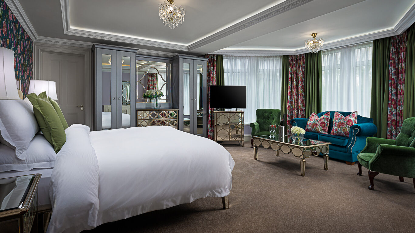 Spacious hotel suite with plush bed, elegant decor, colourful curtains, chandelier, and comfortable seating area.