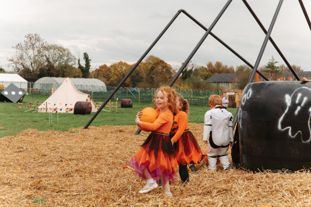 Children in costumes play on a straw-filled field with a tent and decorations, capturing a festive autumn atmosphere.