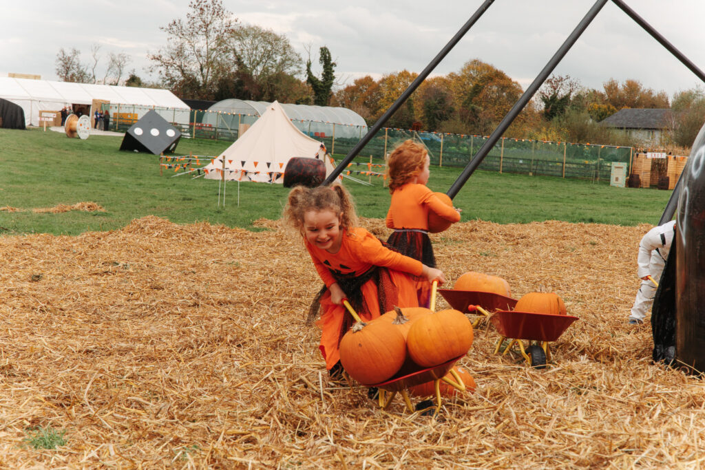 Children in colourful costumes enjoy a pumpkin patch with wheelbarrows, creating festive family memories outdoors.