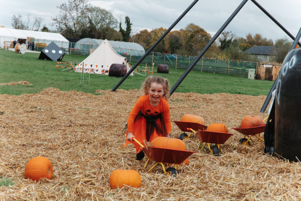 Little girl in orange dress enjoying pumpkin patch with wheelbarrows, playful atmosphere, ideal for family-friendly hotel stays.