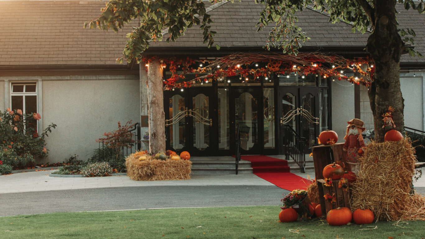 Charming hotel entrance with red carpet, autumn decor, glowing lights, and lush gardens welcoming guests warmly.