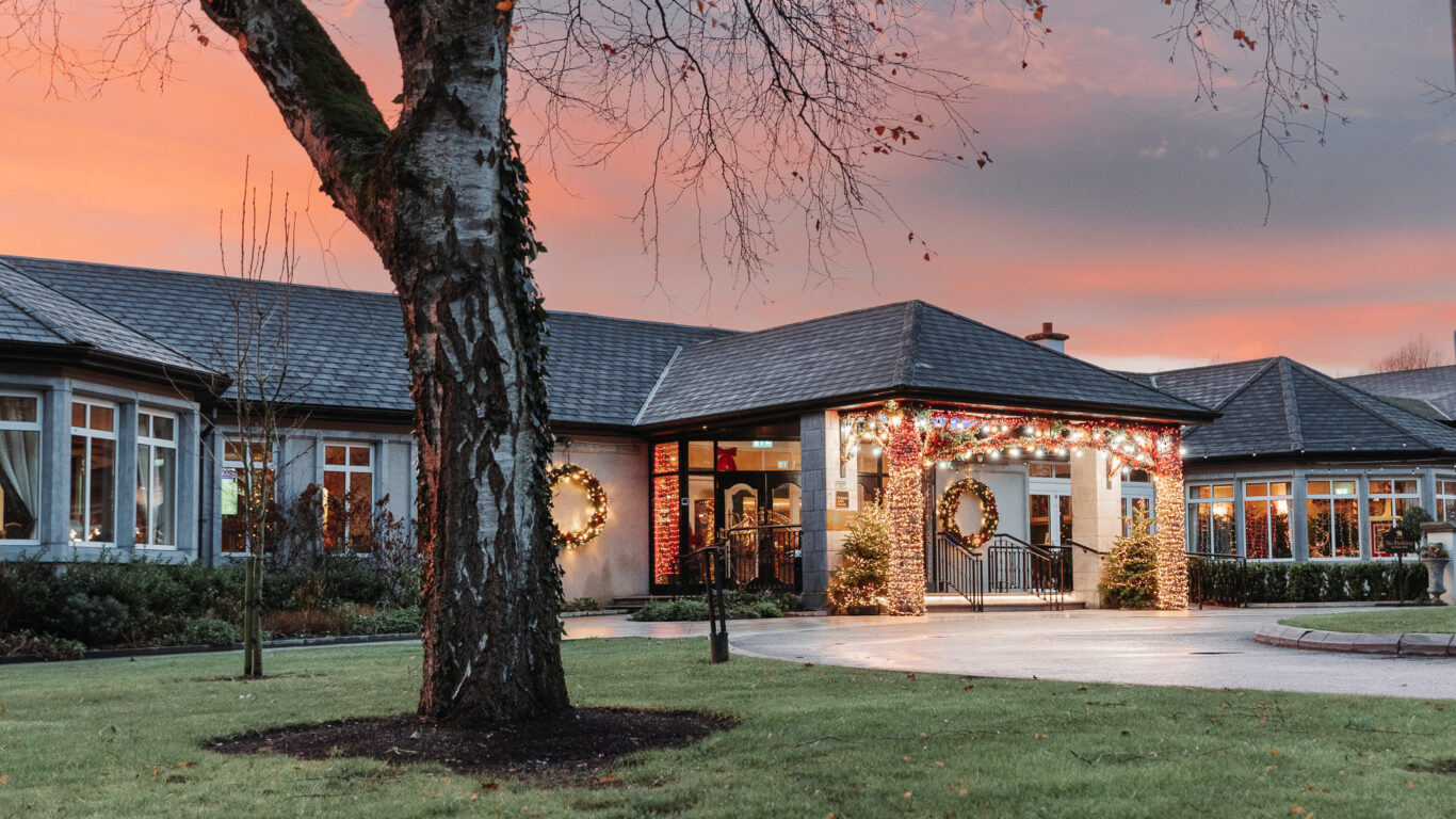 Festive hotel entrance at sunset with twinkling lights and elegant wreaths, surrounded by lush gardens and welcoming ambience.