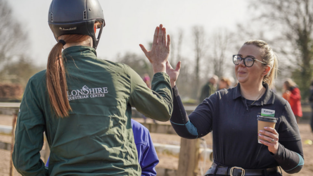 Rider and trainer high-five at scenic equestrian centre, enjoying outdoor horse riding activities.