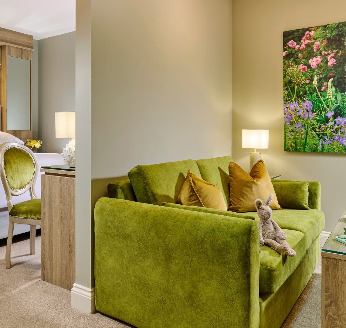 Elegant hotel room with plush green sofa, luxurious bed, floral artwork, and inviting tea service, perfect for relaxation.