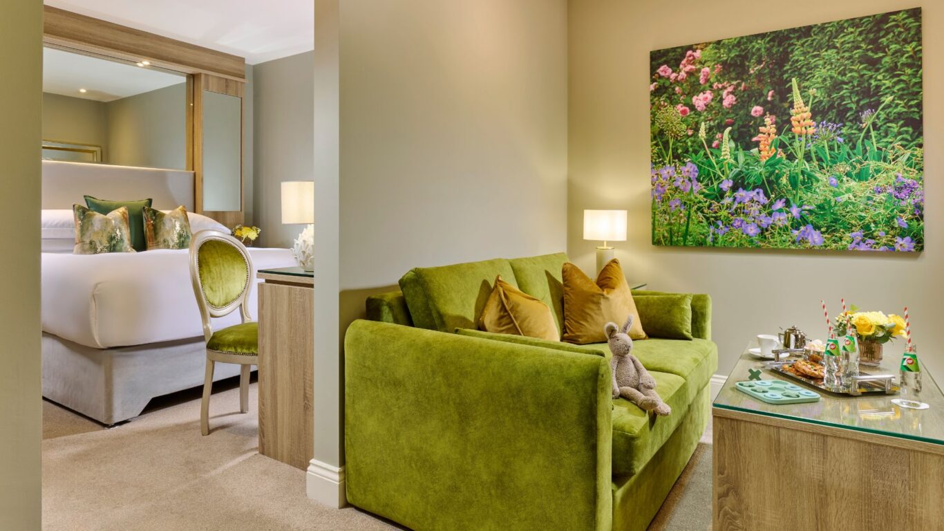 Elegant hotel room with plush green sofa, luxurious bed, floral artwork, and inviting tea service, perfect for relaxation.
