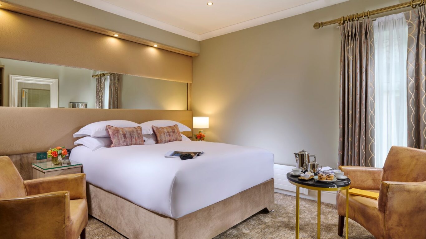 Elegant hotel room with plush bedding, armchairs, and a table set with afternoon tea, offering a cosy and relaxing atmosphere.