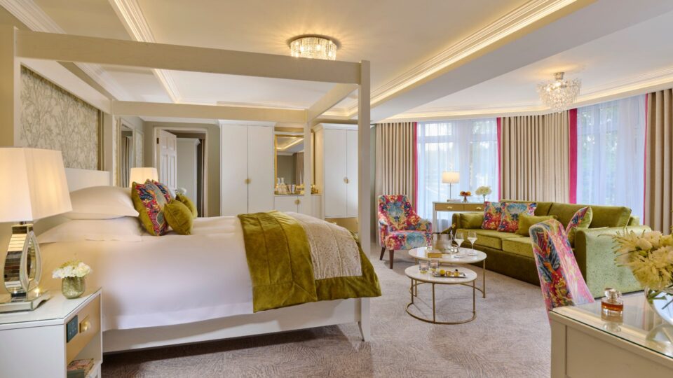 Spacious hotel suite with king canopy bed, vibrant sofa, elegant d&eacute;cor, and natural light, ideal for a relaxing stay.